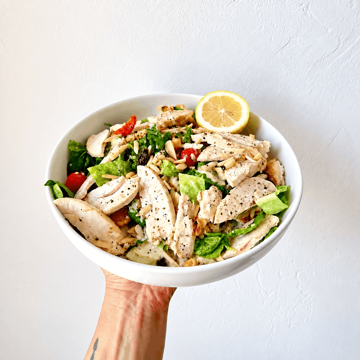 A hand holding a white bowl of chicken salad with greens, tomatoes, almonds, and a lemon slice.